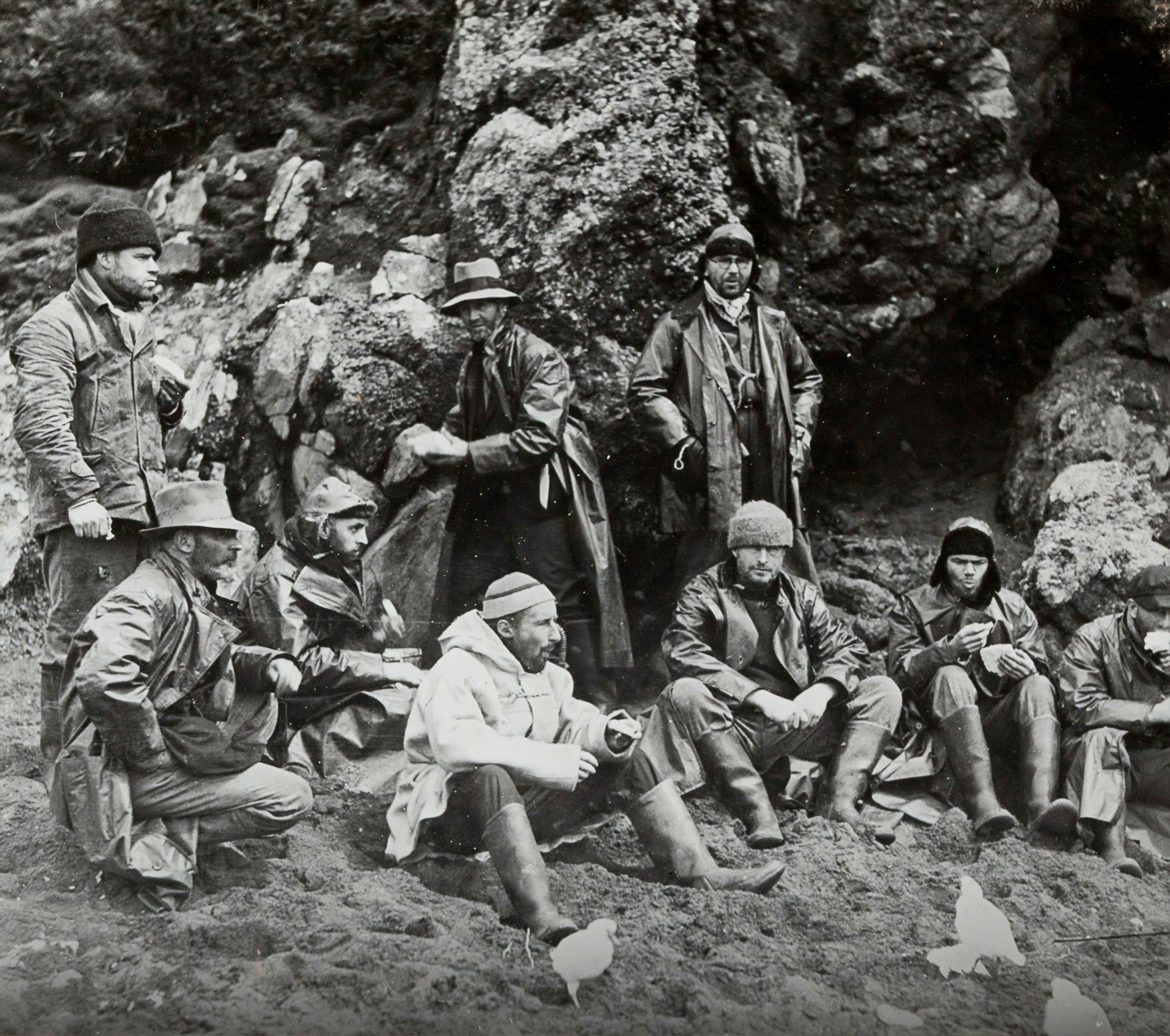 A black-and-white photograph of an expedition team gathered on rocky terrain in Antarctica